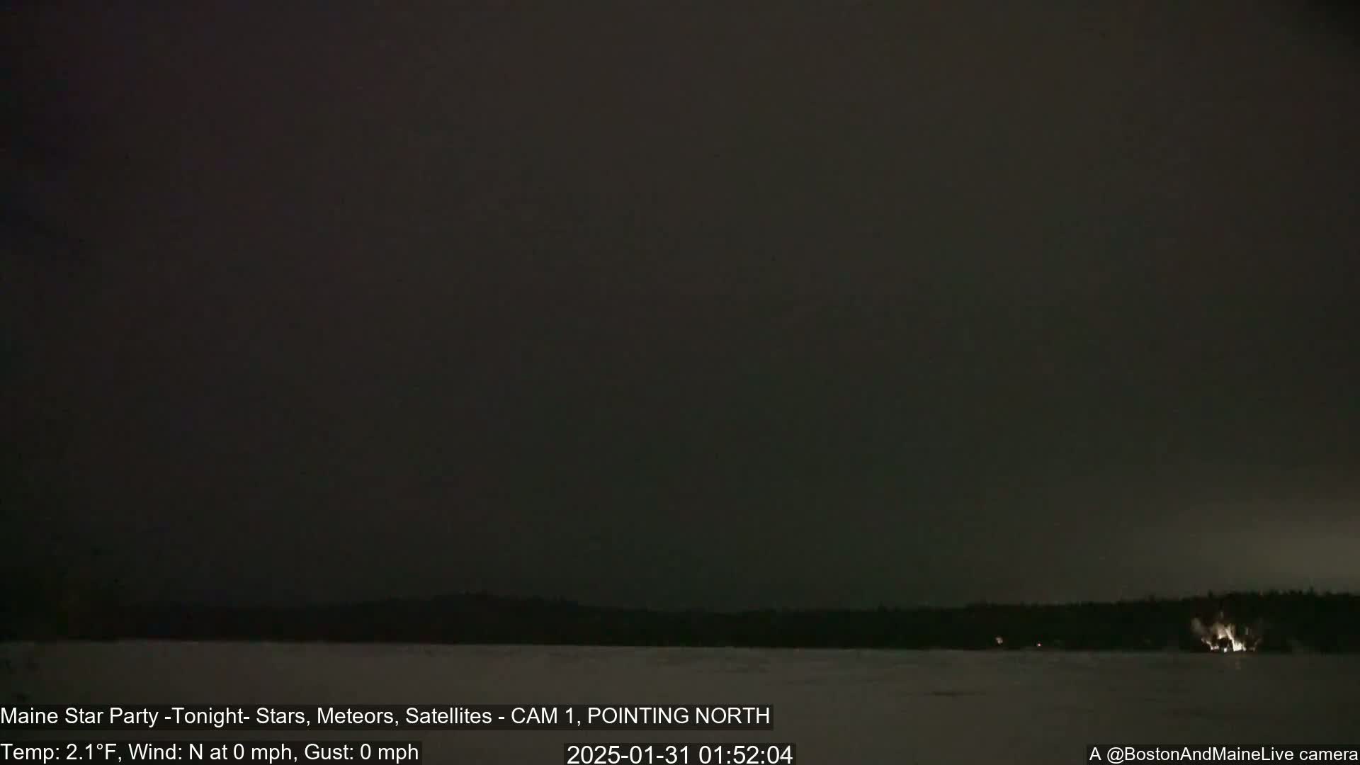 A dark, nighttime scene shows a snow-covered landscape with a treeline in the distance under a mostly clear, dark sky with a temperature of 2.1°F and no wind.