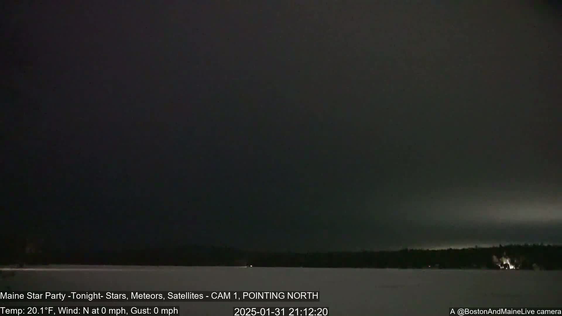 A nighttime view of a snow-covered lake and dark, distant trees under a mostly clear, dark sky with calm winds.