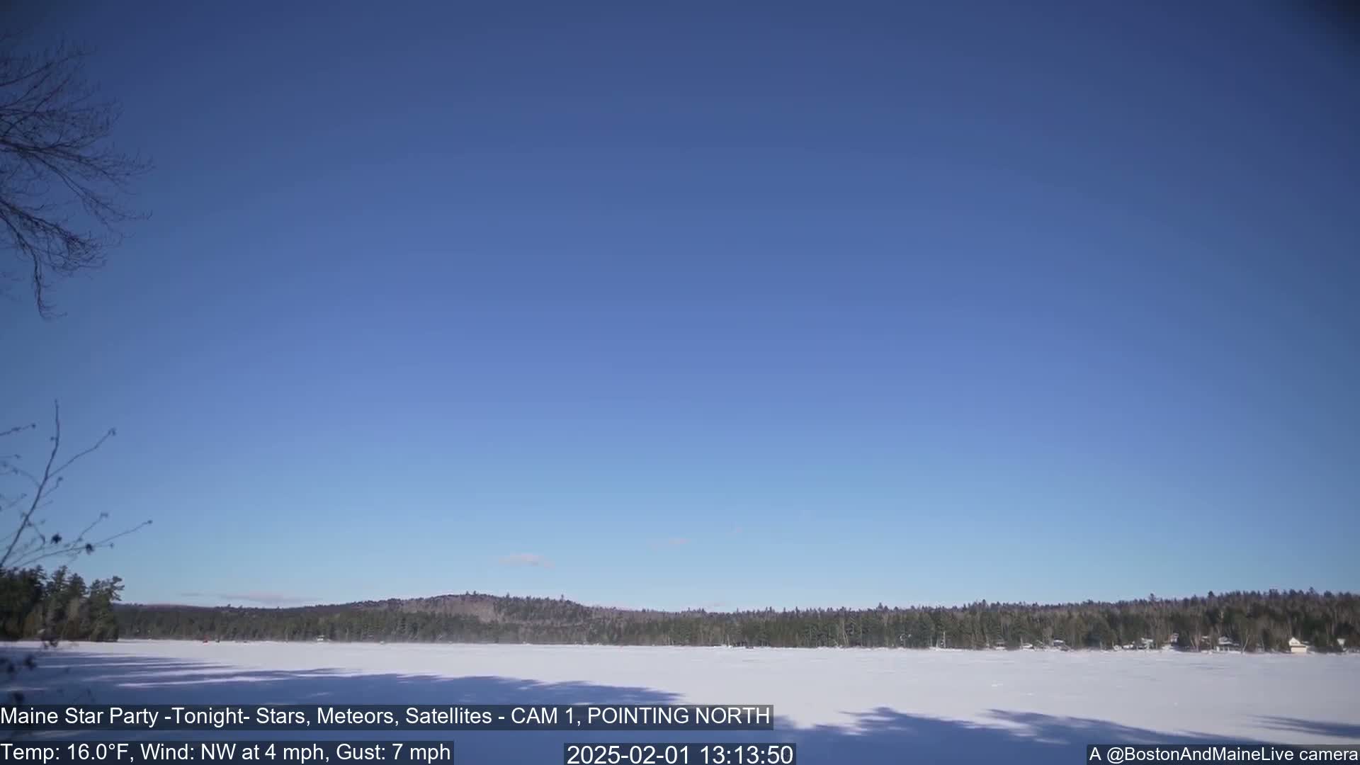 A snow-covered lake is visible, bordered by a dark, tree-lined shore under a clear blue sky, with a light northwest wind.