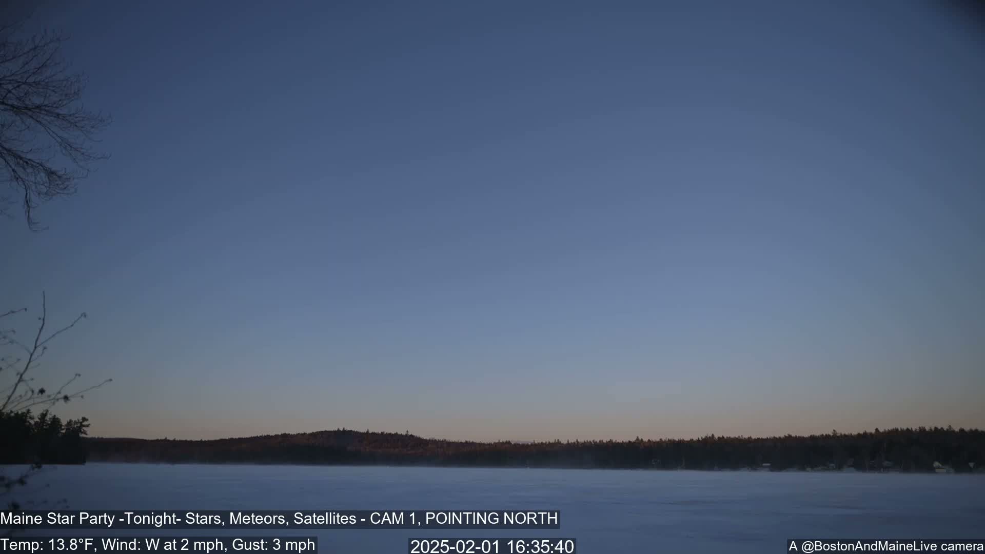 A frozen lake is visible under a twilight sky, with a dark treeline in the distance and bare trees in the foreground, under calm, cool conditions.