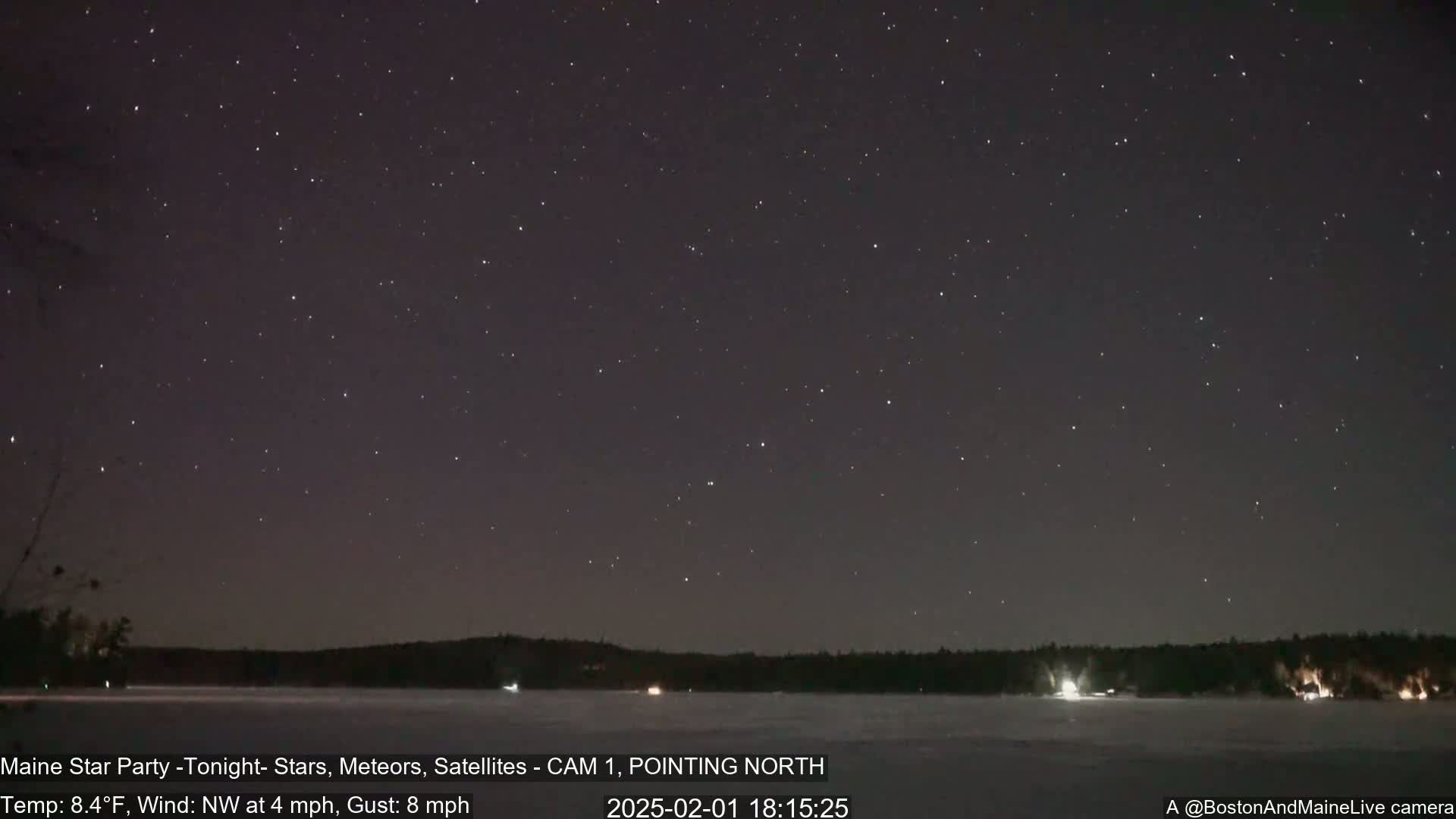 A nighttime view of a star-filled sky over a frozen lake, with a dark treeline in the distance, under a clear sky with a northwest wind.