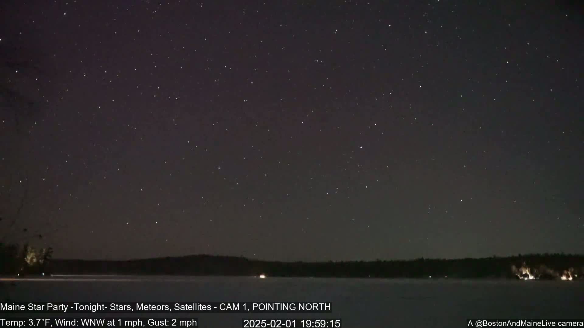 A night sky filled with stars over a dark, frozen lake with a tree line on the horizon, under a cold, clear sky with a light wind.