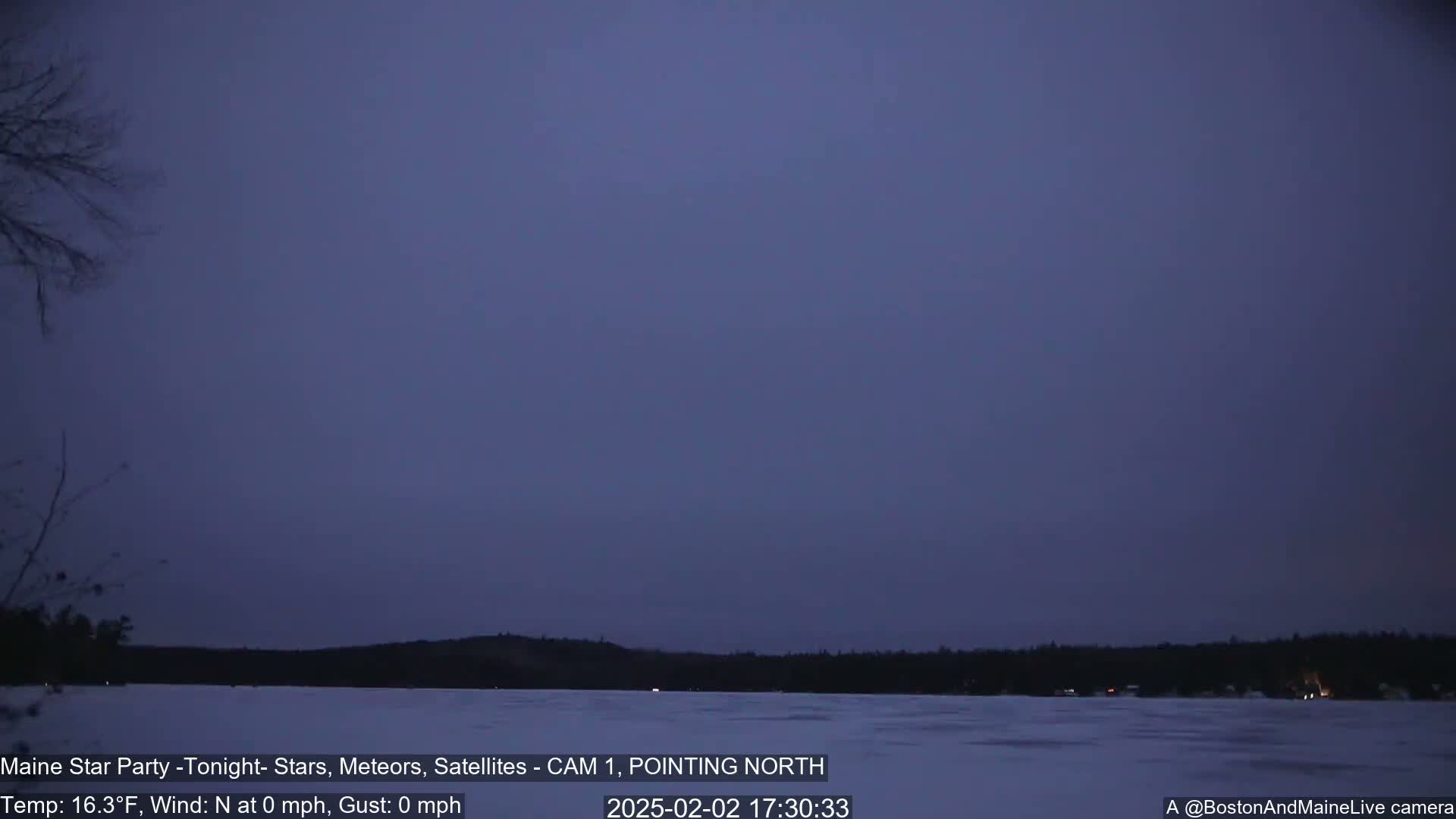 A calm, twilight scene shows a dark, still lake partially covered in ice, with a dark treeline in the background under a dusky purple sky.