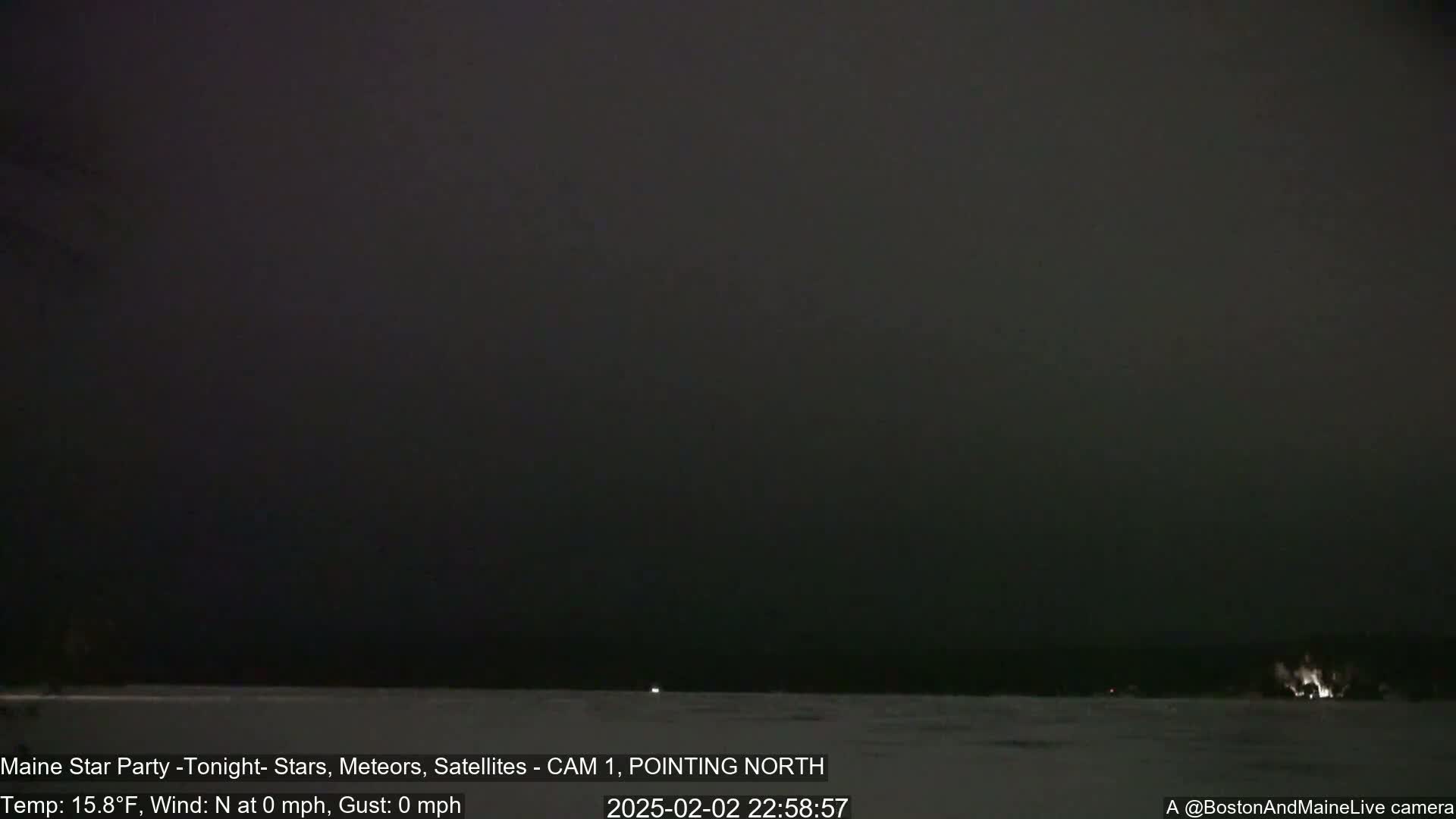 A dark, nighttime scene shows a mostly flat, snow-covered expanse with faint lights in the distance under a dark, overcast sky.