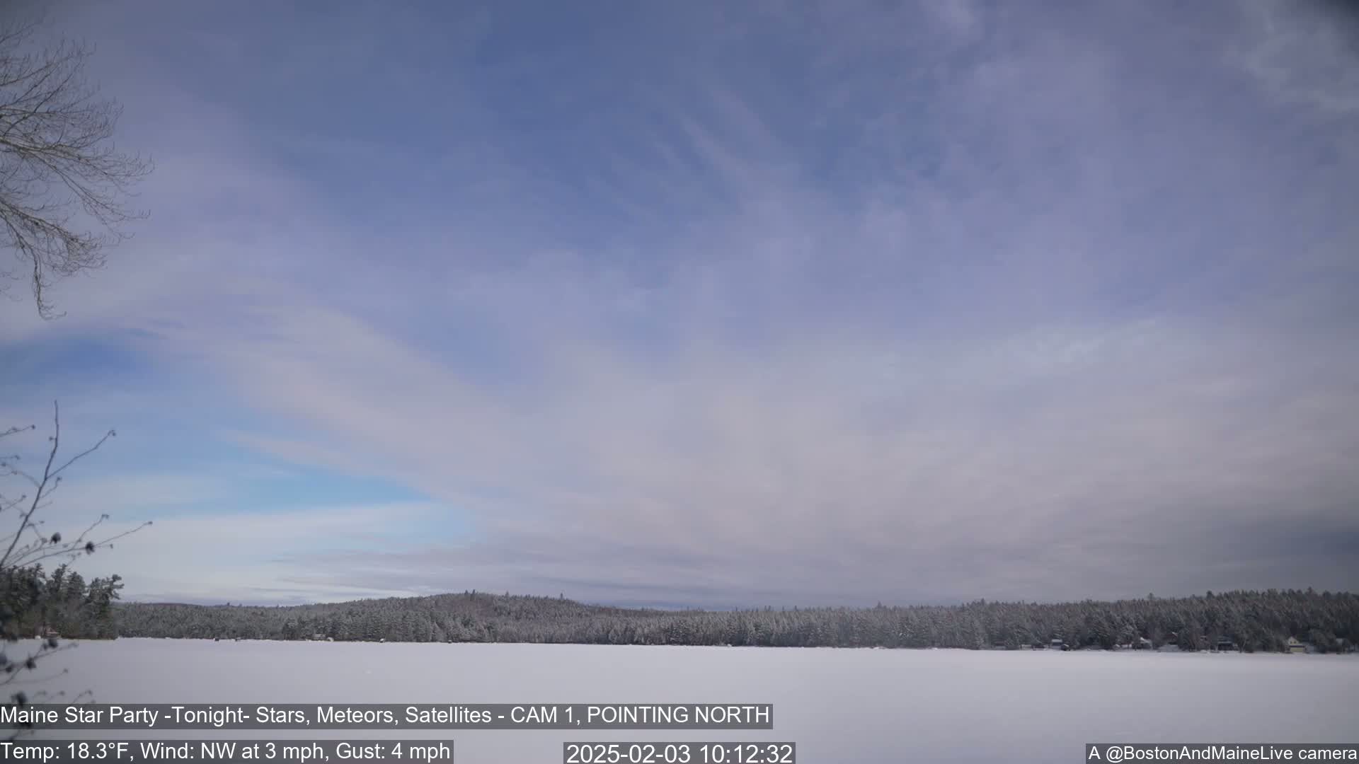 A snow-covered frozen lake is seen under a mostly clear, light-blue sky with thin, wispy clouds; a snow-dusted treeline is visible in the background.