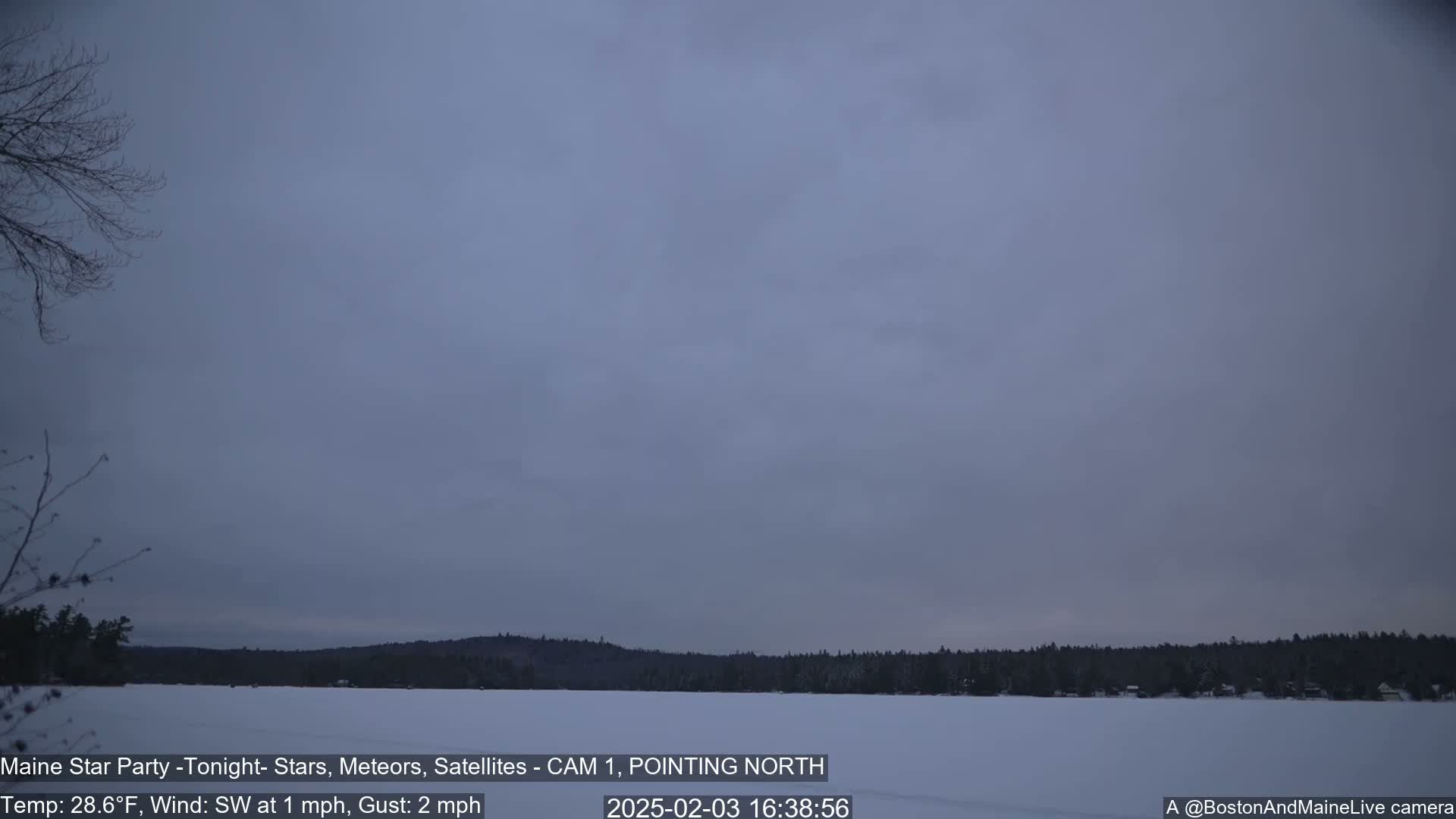 A snow-covered frozen lake is visible, with a dark, overcast sky and a treeline in the background under a mostly cloudy sky with a light southwest wind.