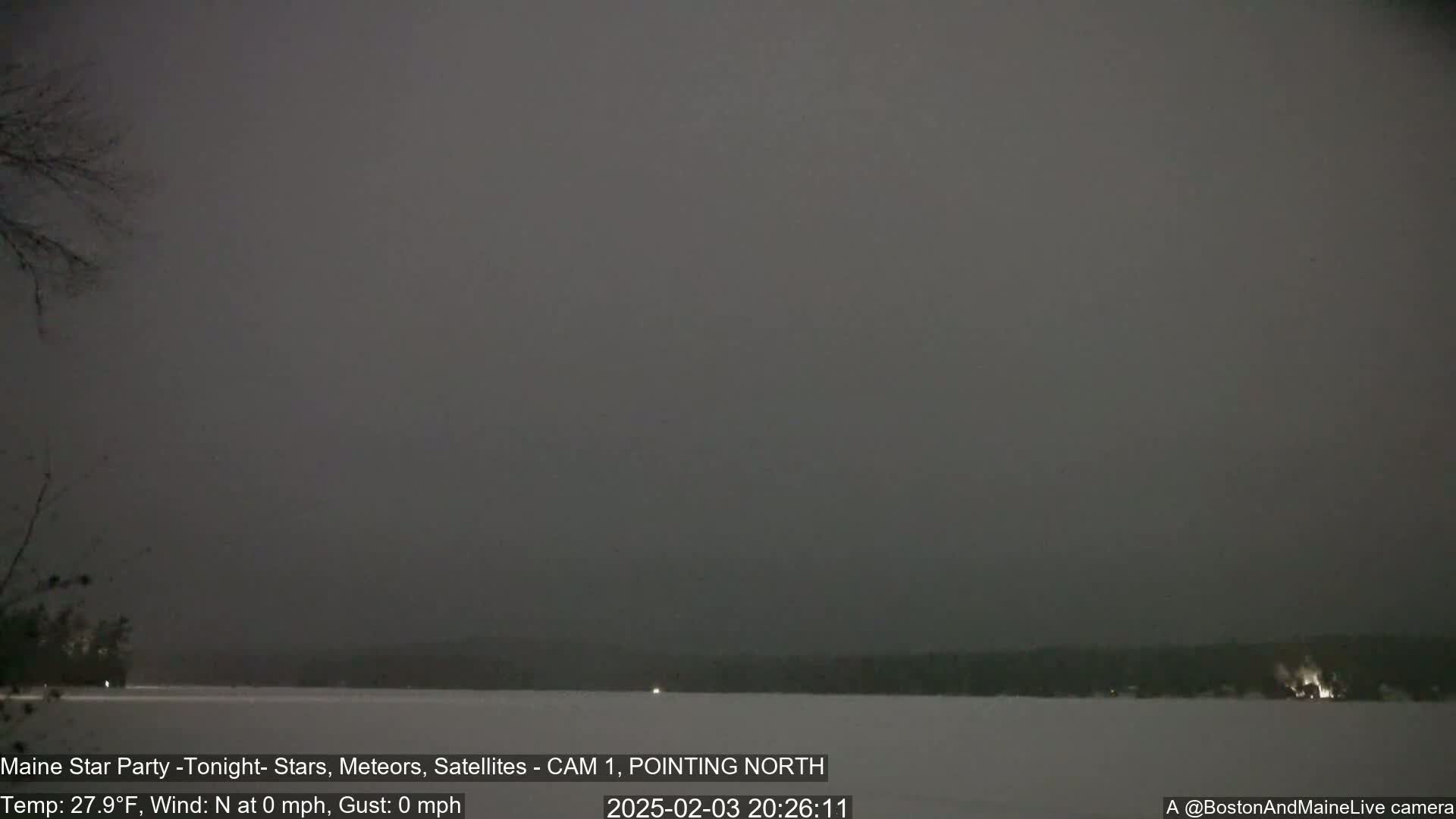 A dark, snowy landscape is visible at night, showing a body of water covered in snow and a treeline in the distance under an overcast sky.