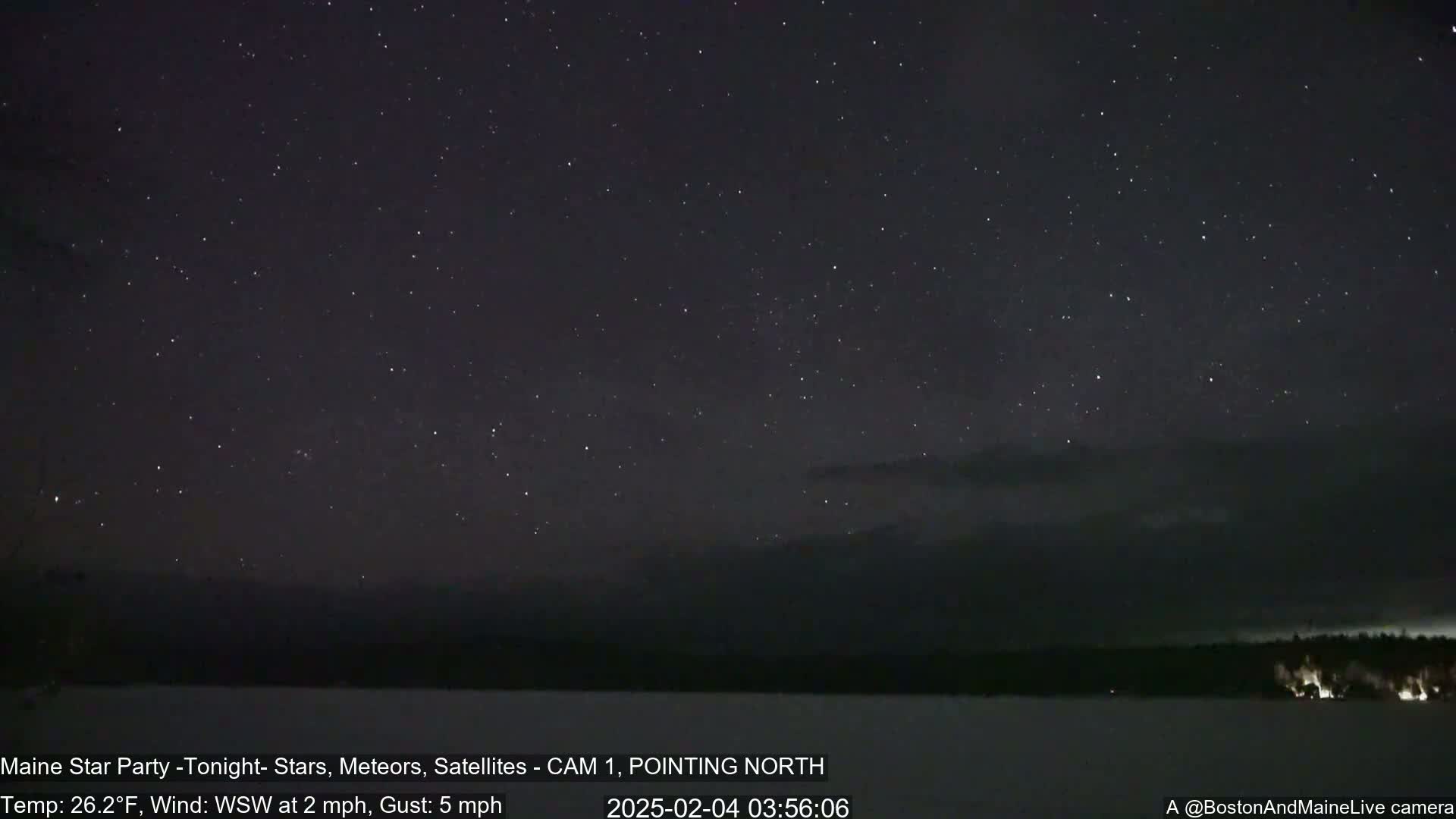 A nighttime scene shows a dark sky full of stars over a snow-covered landscape with a dark treeline, under a cold, windy sky.