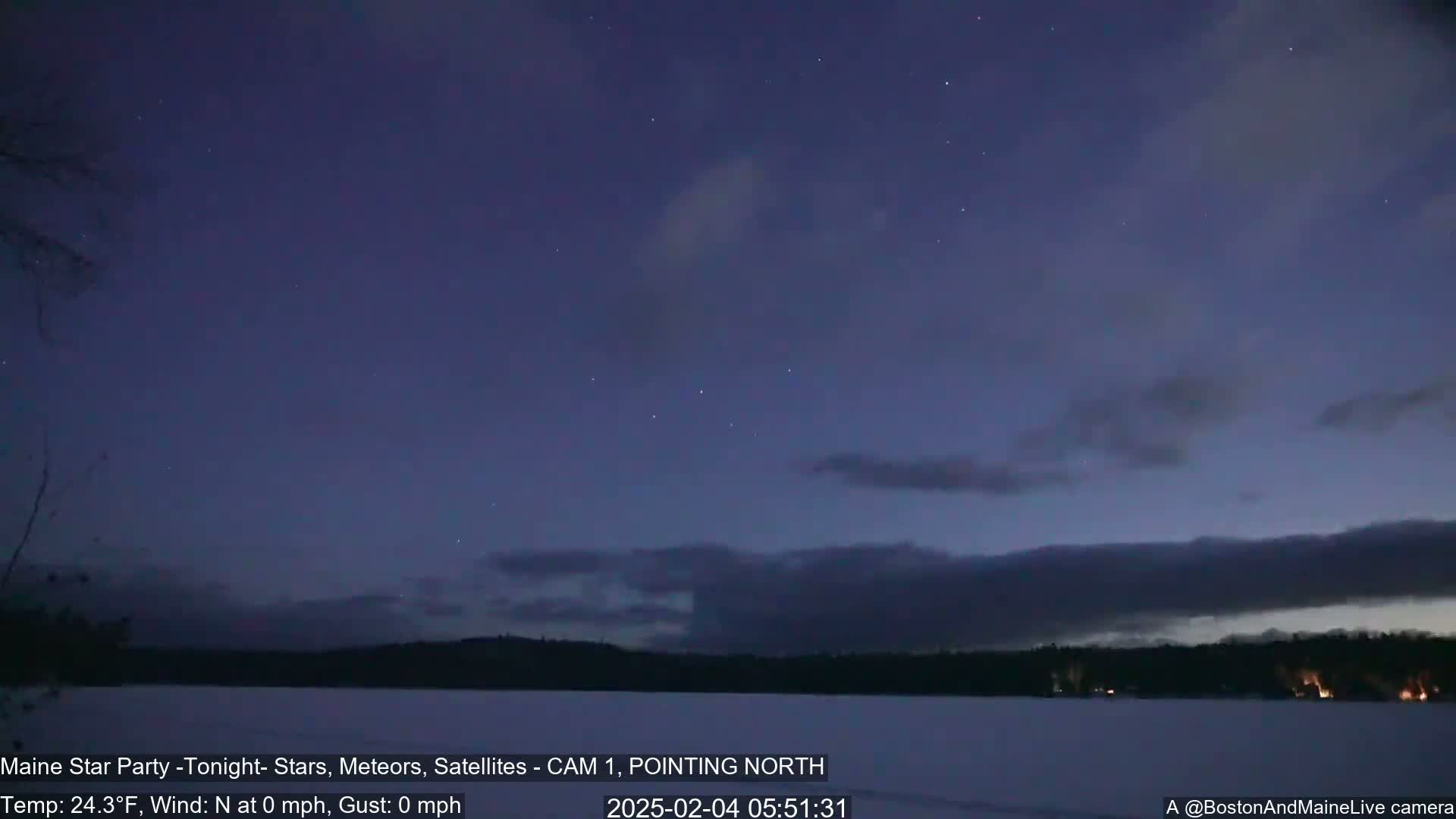 A calm, dark night sky with a few stars and thin clouds over a snow-covered lake and dark treeline.