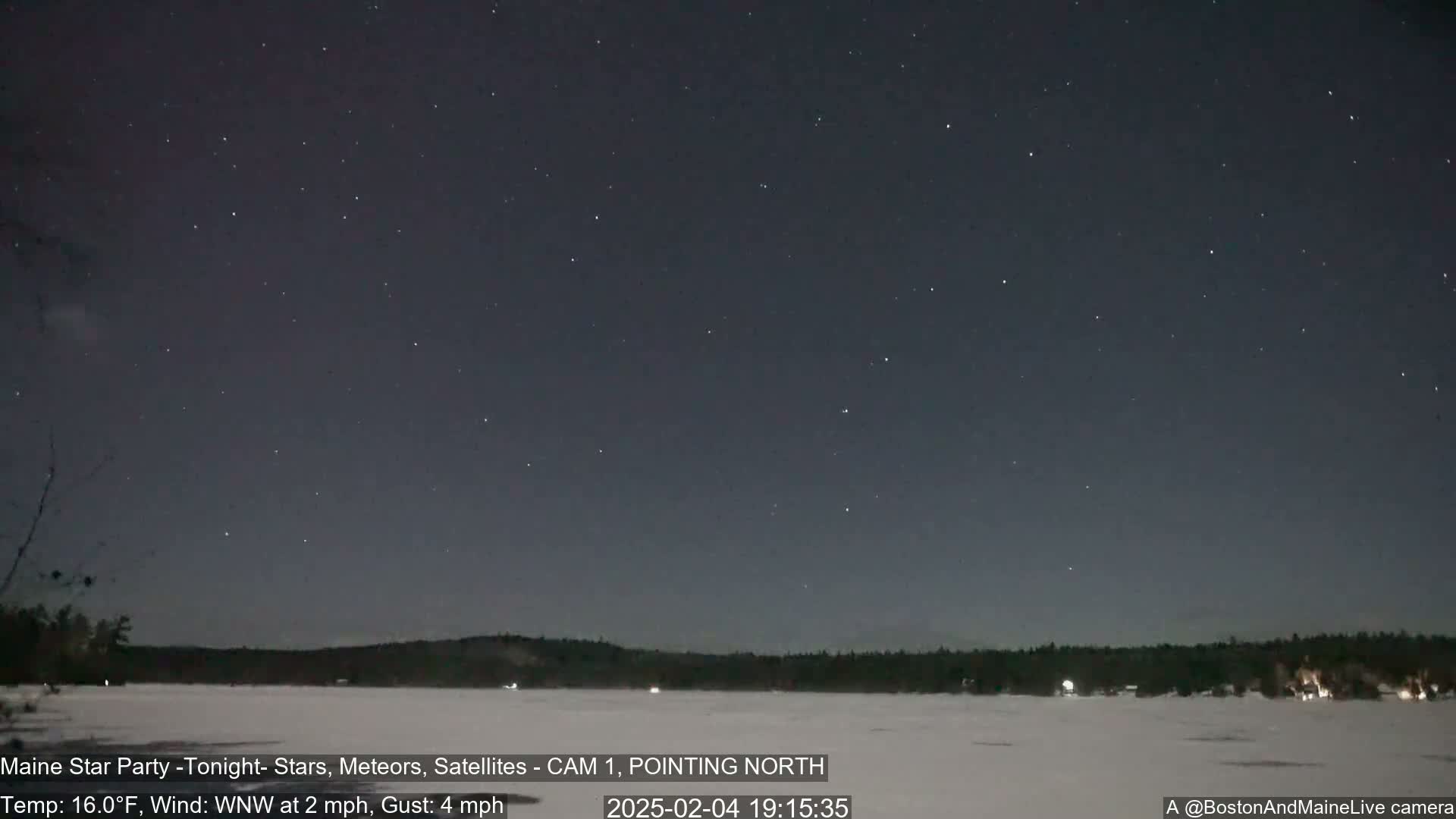 A nighttime view of a snow-covered frozen lake, with a dark sky full of stars and a tree line in the distance, under a clear, cold sky with a light wind.