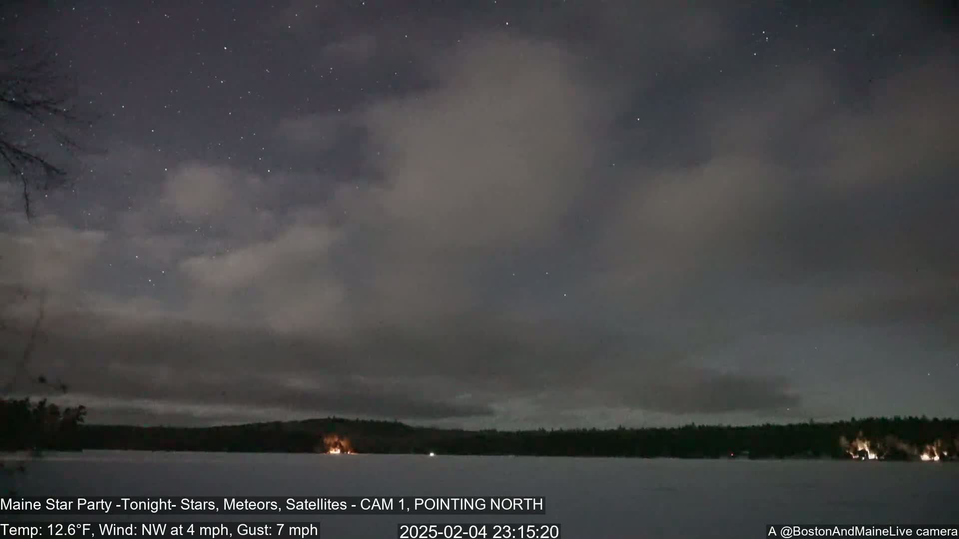 A mostly cloudy night sky with some stars visible over a frozen lake and a dark treeline, with a northwest wind at 4 mph and gusts up to 7 mph.