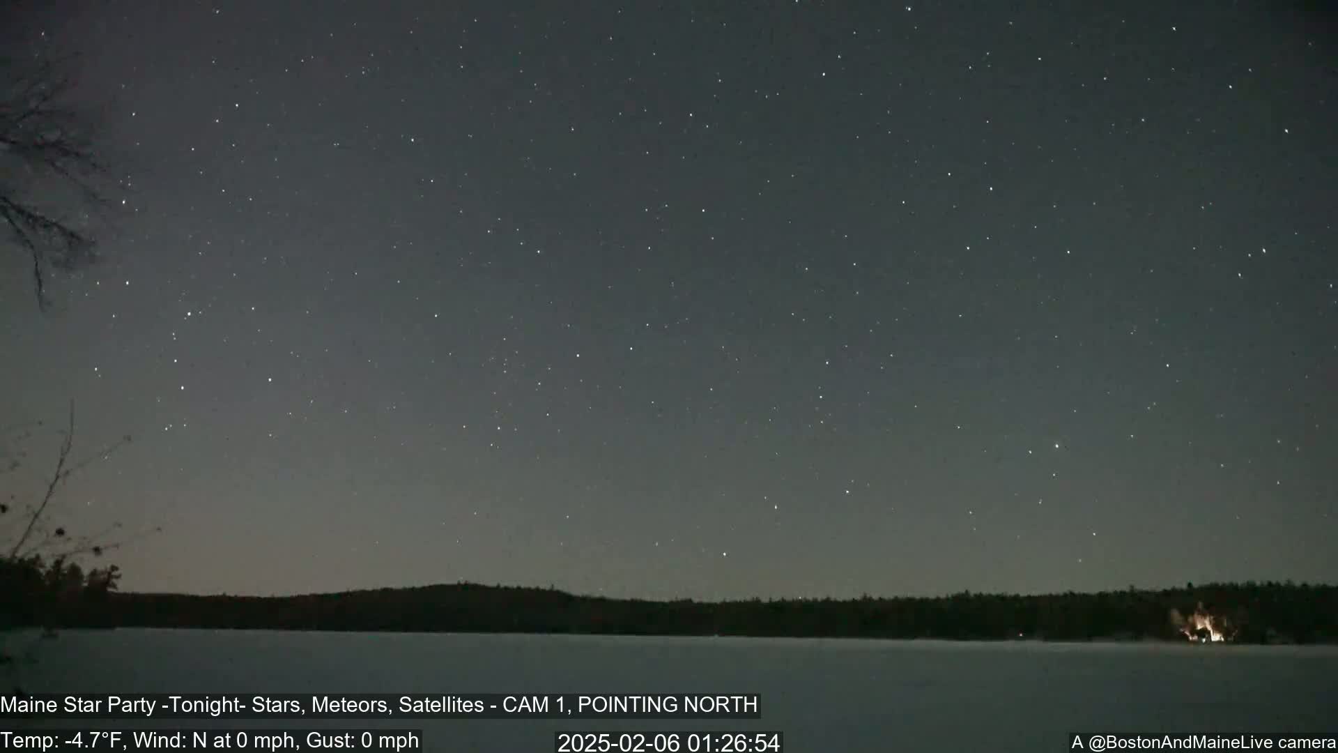 A nighttime long shot shows a dark, clear sky full of stars over a frozen lake with a dark treeline in the background; there is a small fire visible on the far shore.