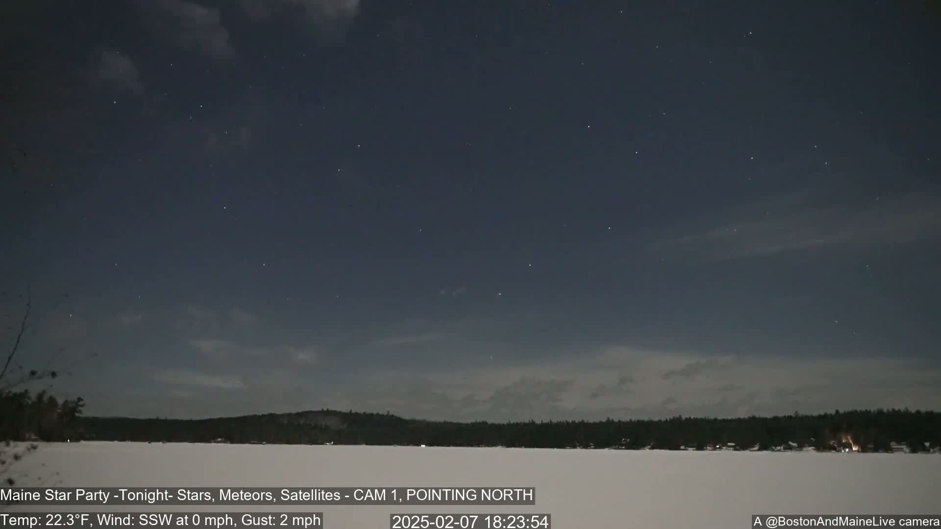 A nighttime long shot shows a starry sky over a snow-covered frozen lake and dark trees under mostly clear, slightly cloudy conditions.