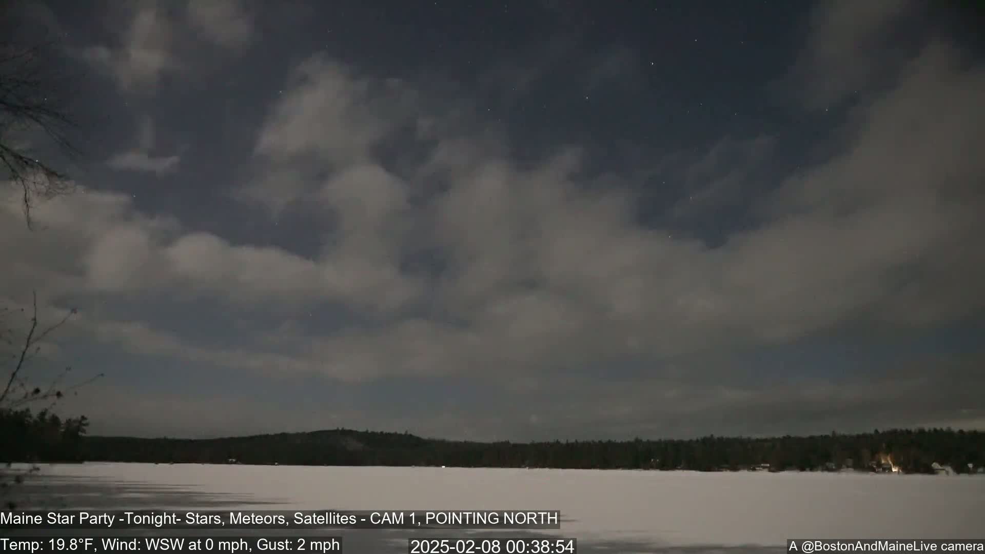 A partly cloudy night sky over a frozen lake and snow-covered shoreline under a nearly full moon.