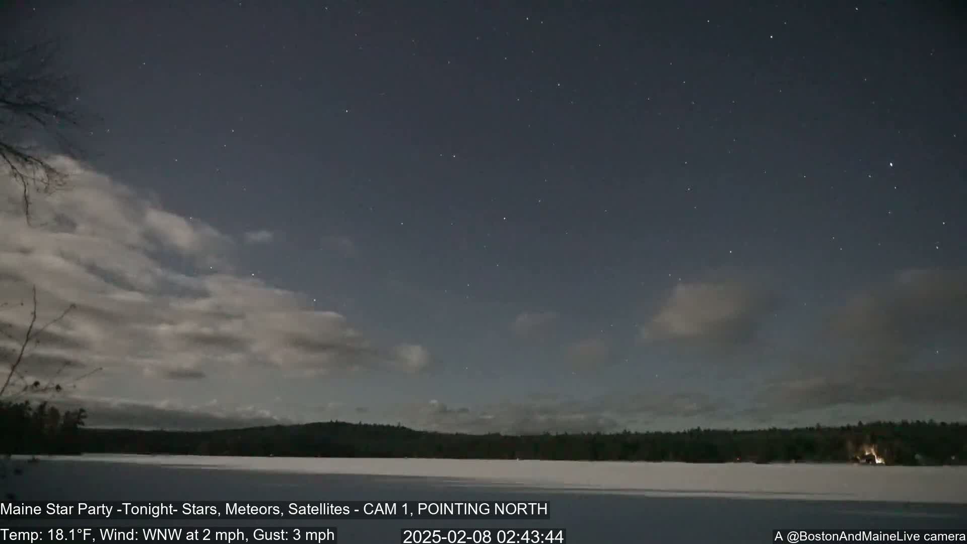 A mostly cloudy night sky with visible stars over a snow-covered frozen lake and dark treeline, with a light wind.