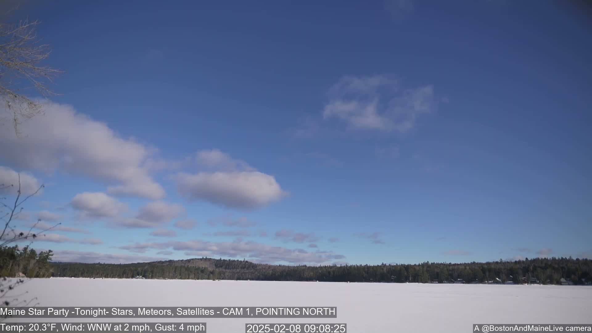 A mostly clear, blue sky with scattered clouds over a snow-covered frozen lake and a distant, dark treeline.