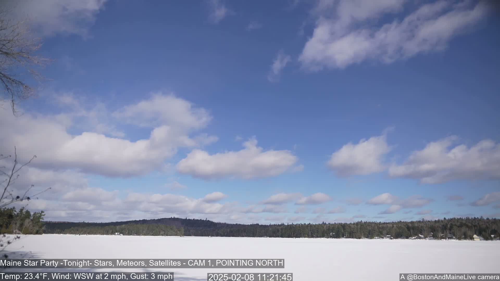 A mostly sunny, cold day with a few scattered clouds over a snow-covered lake and a distant tree line.