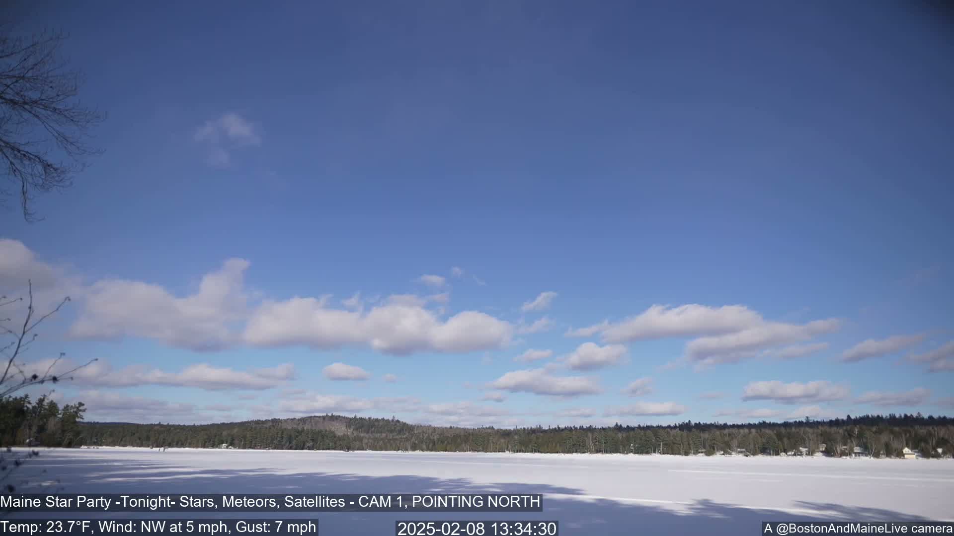 A snow-covered lake is surrounded by a dark, treeline under a mostly clear, blue sky with scattered cumulus clouds.