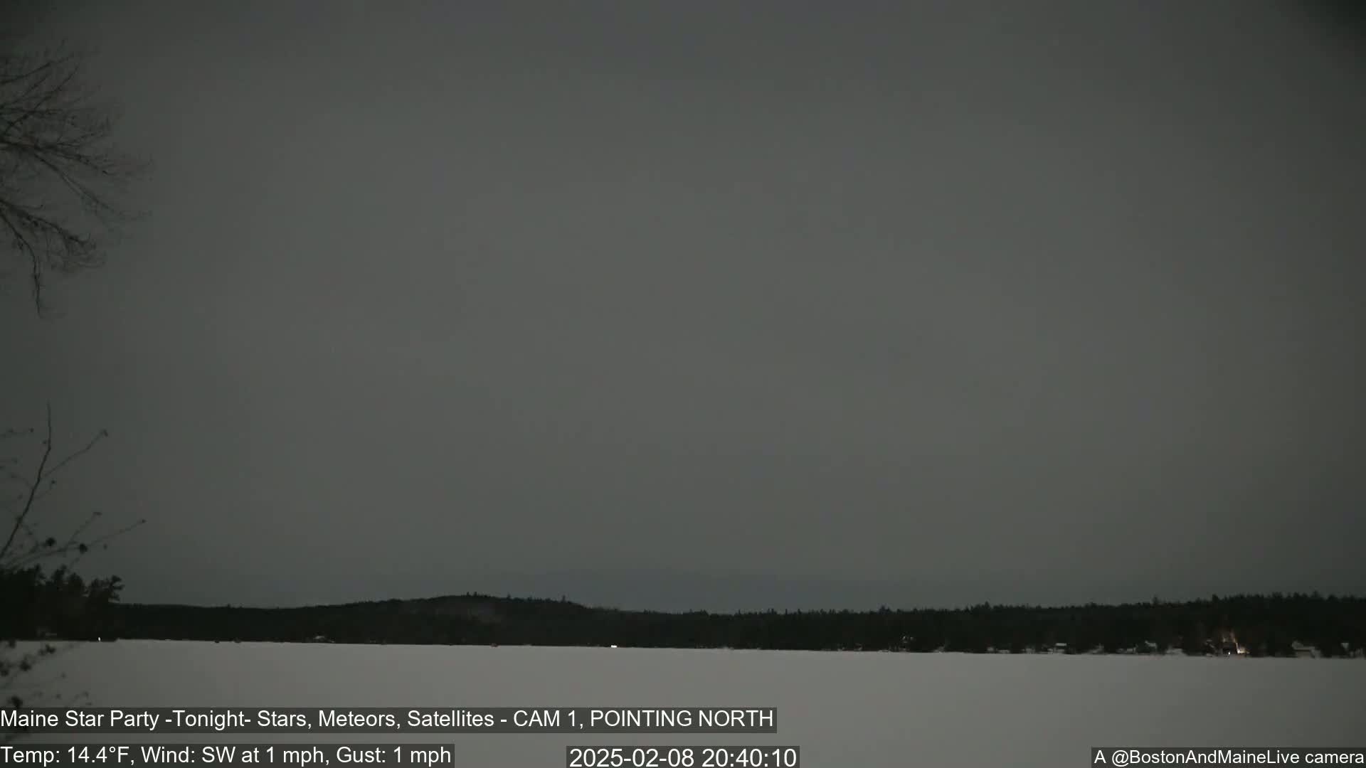 A nighttime long shot shows a snow-covered landscape with a dark gray sky, a calm lake, and dark silhouettes of trees in the background under a 14.4°F temperature with a southwest wind of 1 mph.