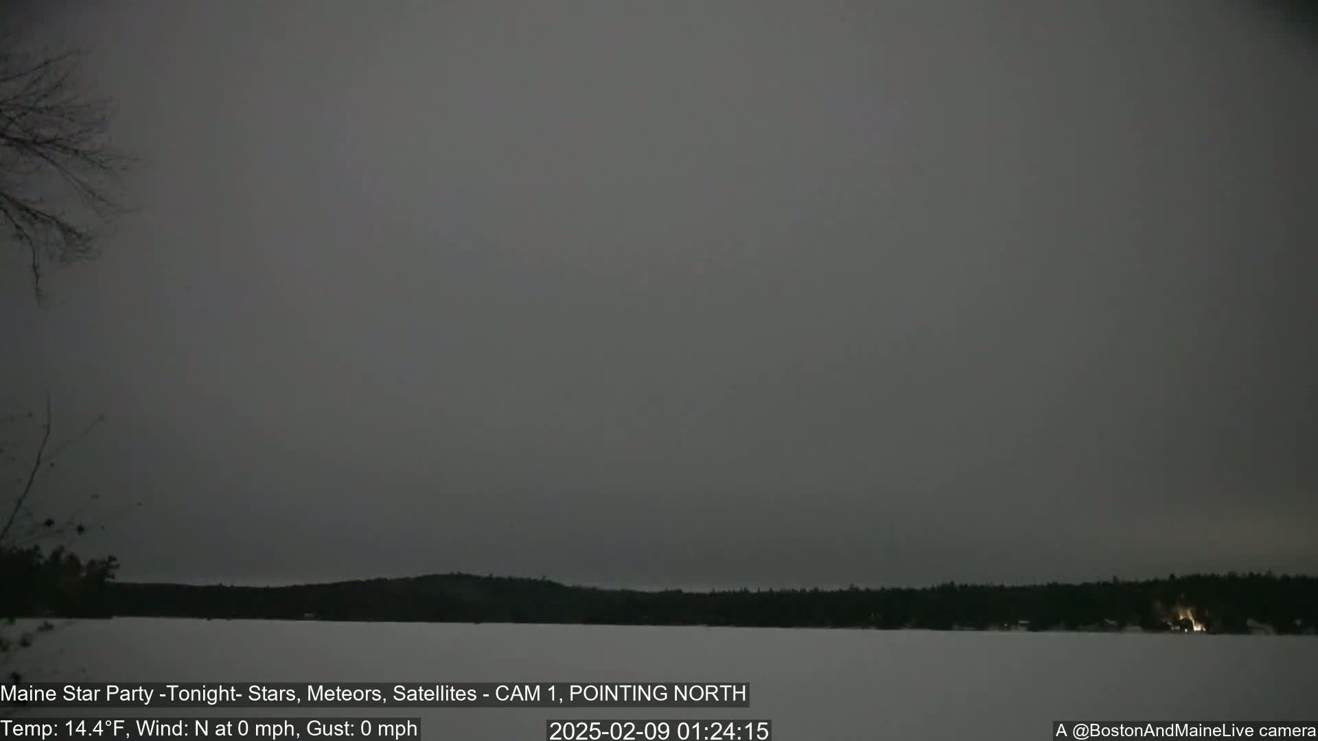 A dark, overcast night scene shows a snow-covered lake and distant trees under a gray sky with no wind.