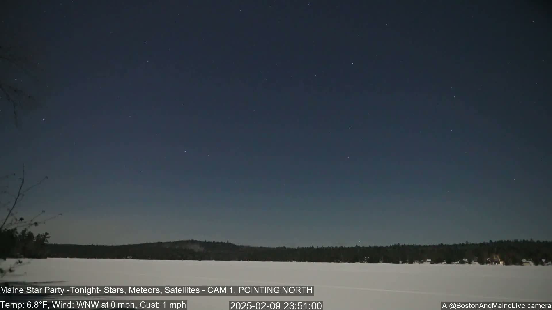 A clear night sky with numerous stars is visible over a snow-covered frozen lake and dark treeline, under calm, cold conditions.