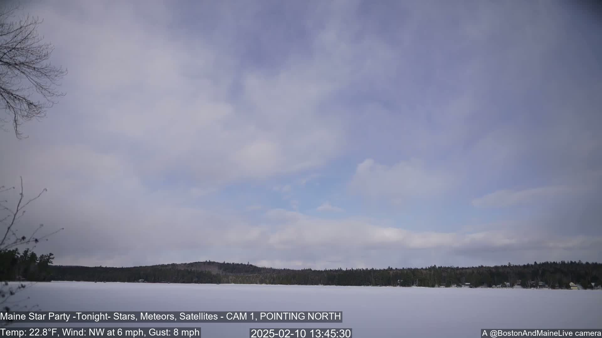 A snow-covered lake is visible beneath a mostly cloudy sky, with a dark treeline in the background under a light wind.