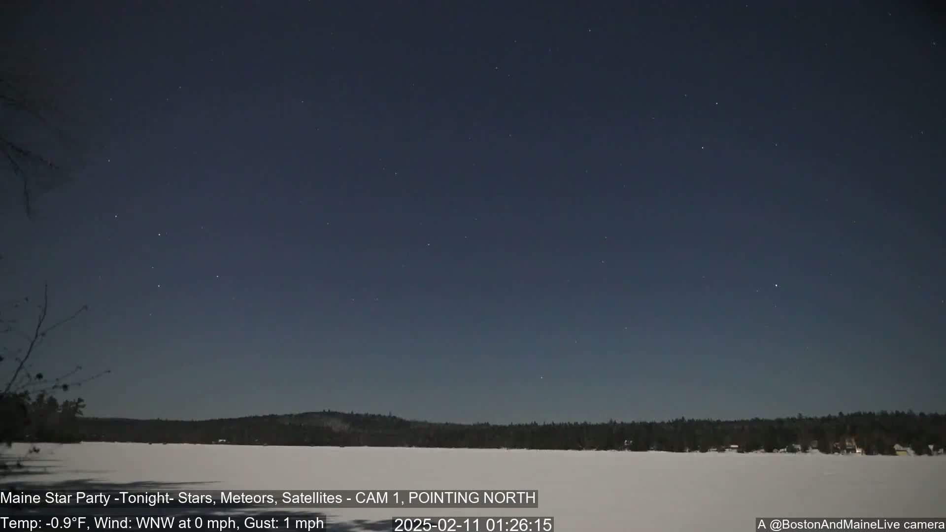 A nighttime scene shows a clear, star-filled sky over a snow-covered landscape with a dark tree line in the distance under calm, cold conditions.