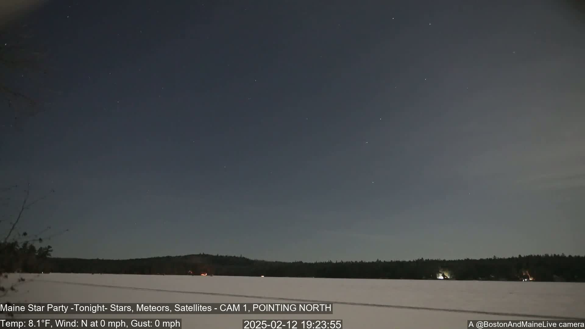 A clear night sky with many stars is over a snow-covered, frozen body of water and a dark treeline.