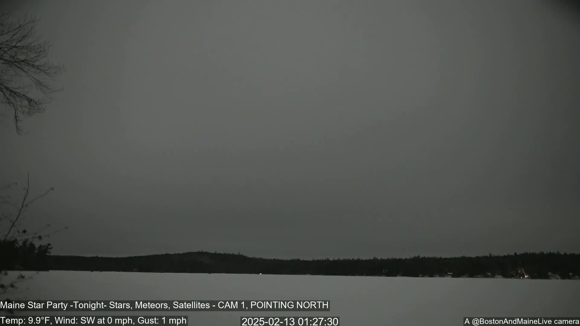 A dark, overcast sky hangs over a snow-covered lake and dark trees at night.