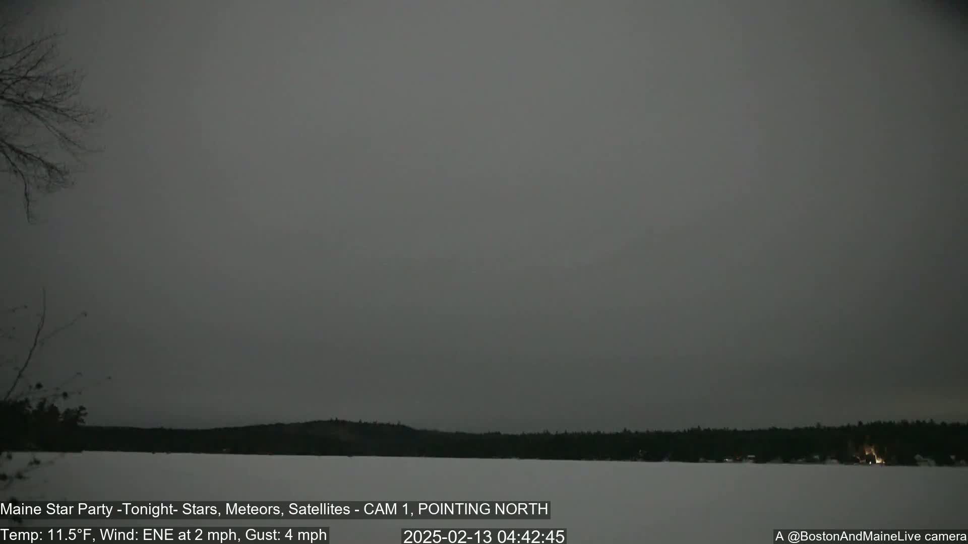 A snow-covered landscape with a dark, overcast sky shows a treeline across a frozen body of water under a gray sky.