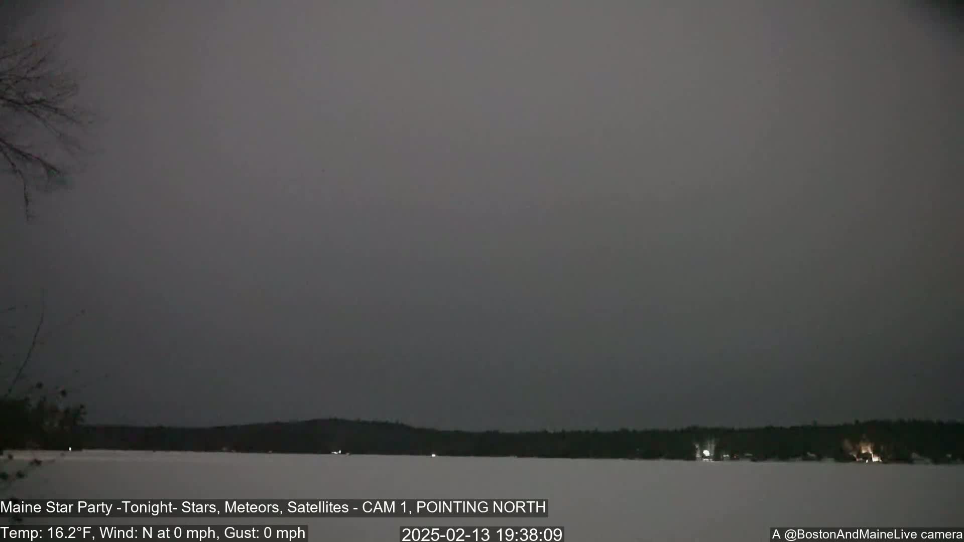 A dark, overcast night scene shows a snow-covered lake with a tree line in the distance, under calm, windless conditions.