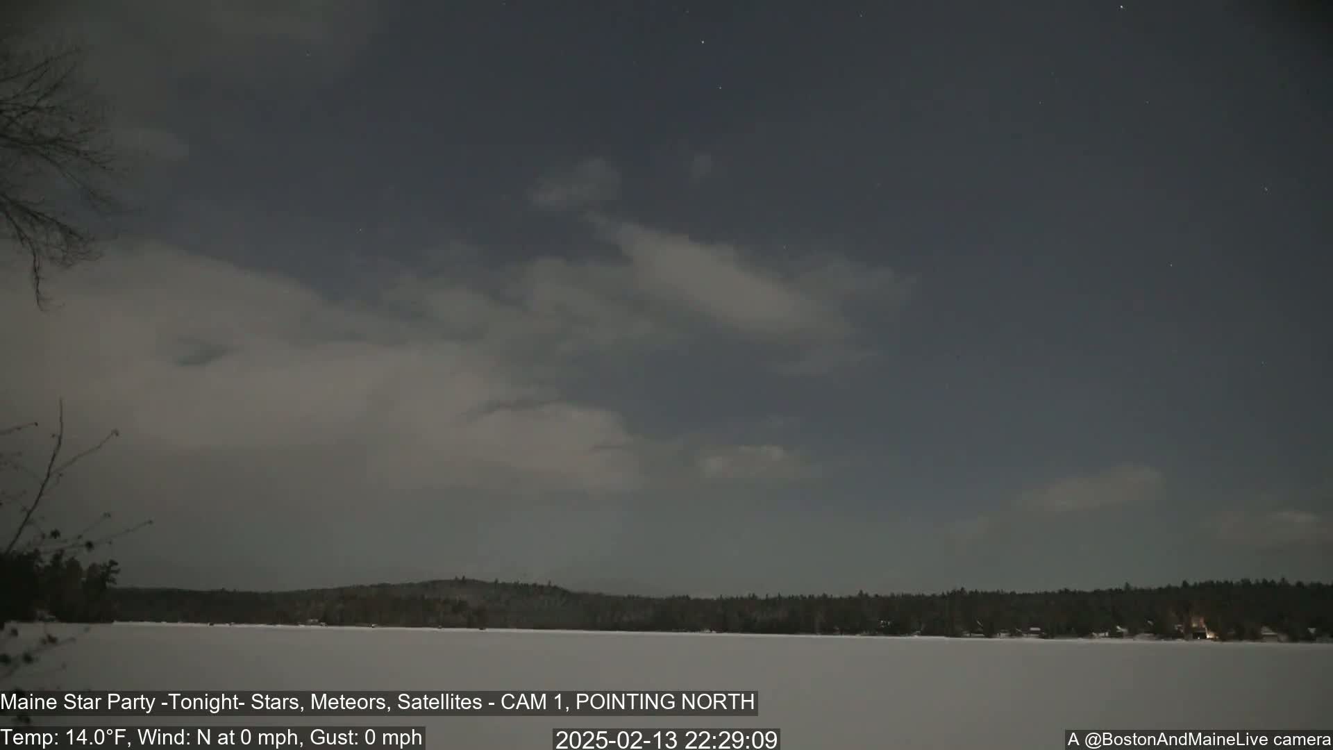 A mostly cloudy night sky over a snow-covered frozen lake and dark, tree-lined shore.