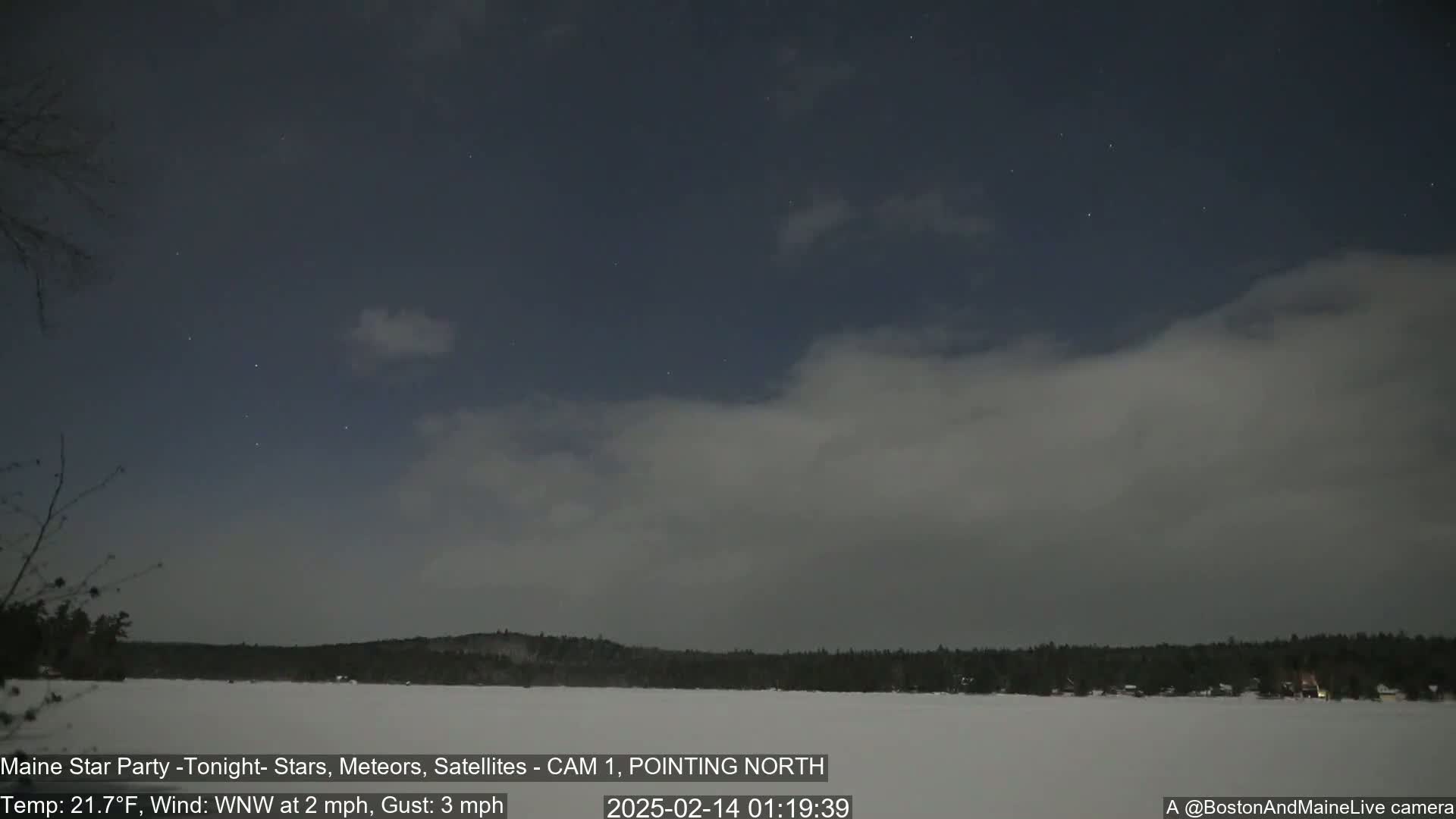 A nighttime view of a snow-covered lake and tree line under a partly cloudy sky with visible stars.