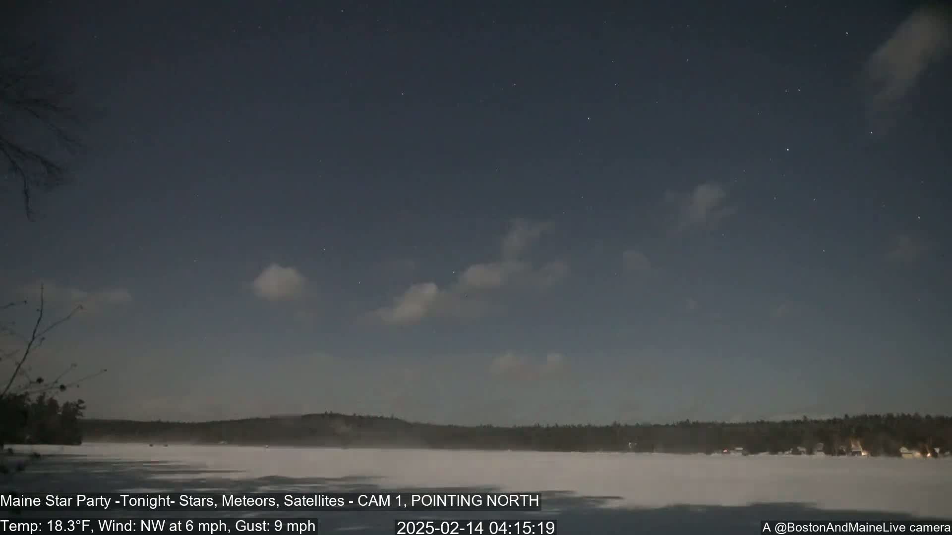 A nighttime view of a frozen lake and snow-covered shoreline under a partly cloudy sky with a few stars visible.