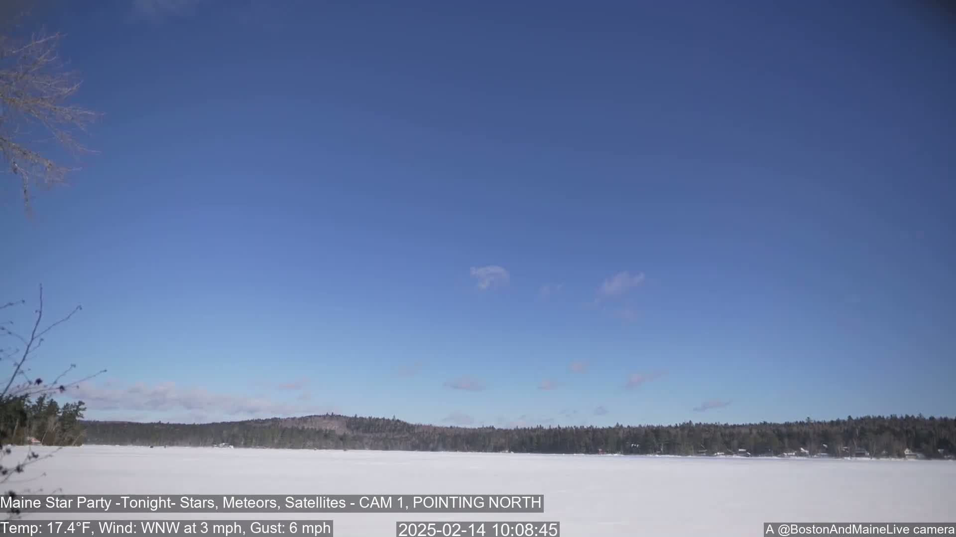 A snow-covered lake is seen beneath a mostly clear blue sky with a few wispy clouds, and a dark treeline is visible in the distance.