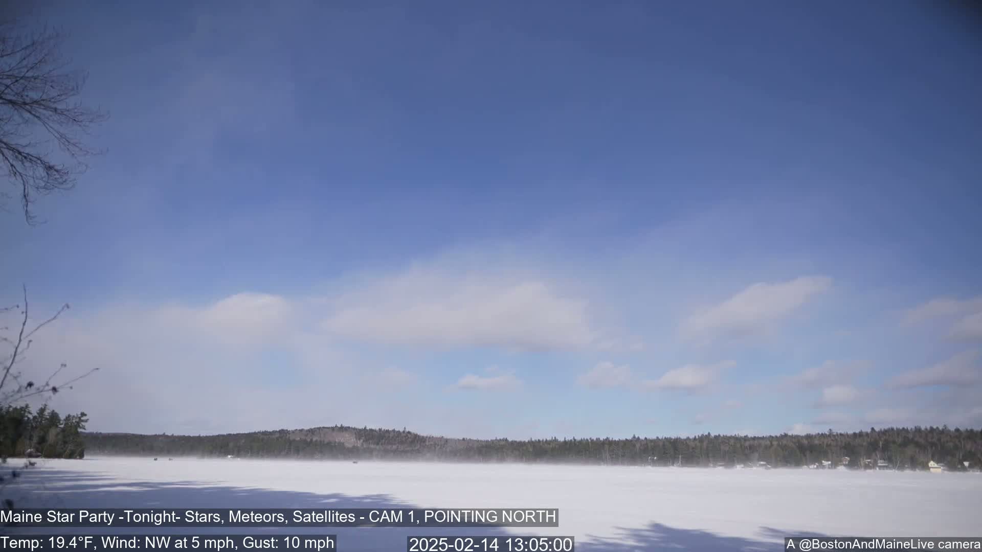 A snow-covered lake stretches to a treeline under a mostly clear, light blue sky with a few wispy clouds, and a light wind is blowing.