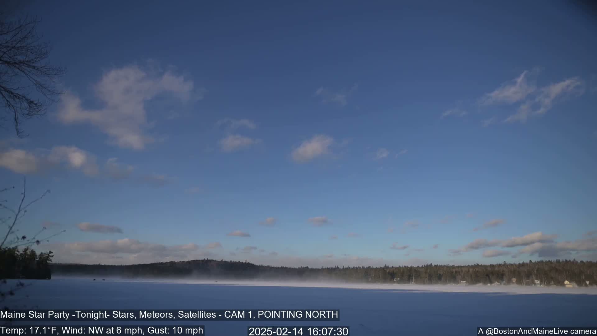 A snow-covered lake is visible beneath a mostly clear, light-blue sky with a few scattered clouds, and a line of dark trees stretches across the horizon.