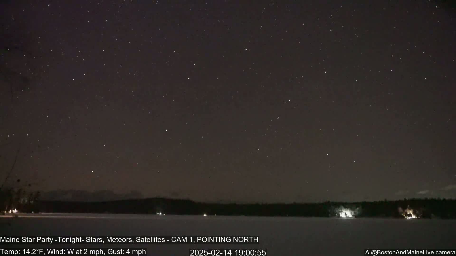 A clear night sky filled with stars is visible over a snow-covered lake and dark treeline, under a cold, windy condition.