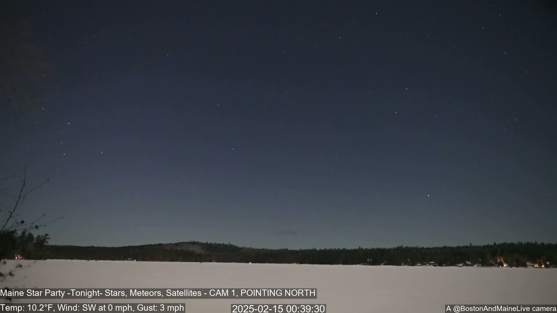 A night sky filled with stars overlooks a snow-covered frozen lake and dark treeline, under calm, cold conditions.