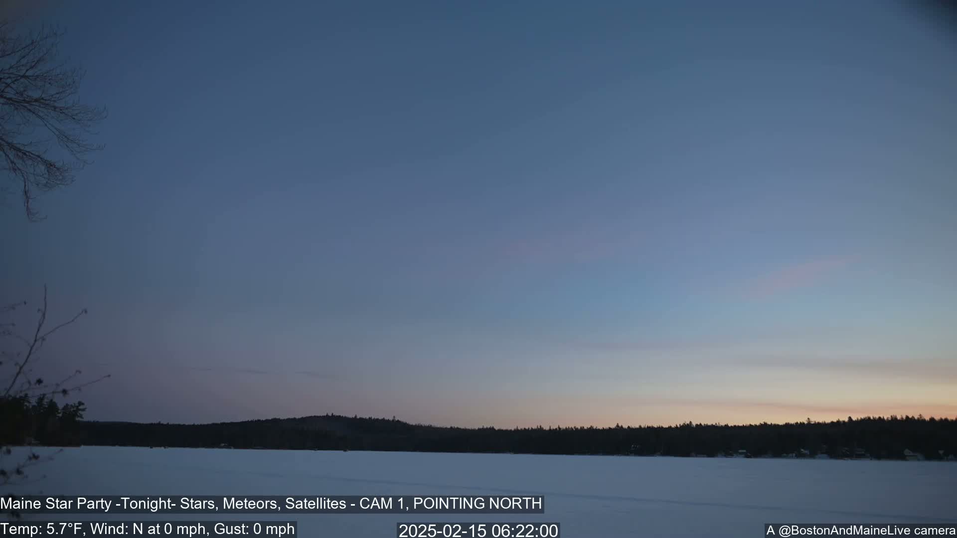 A calm, pre-dawn sky over a snow-covered lake and dark treeline.