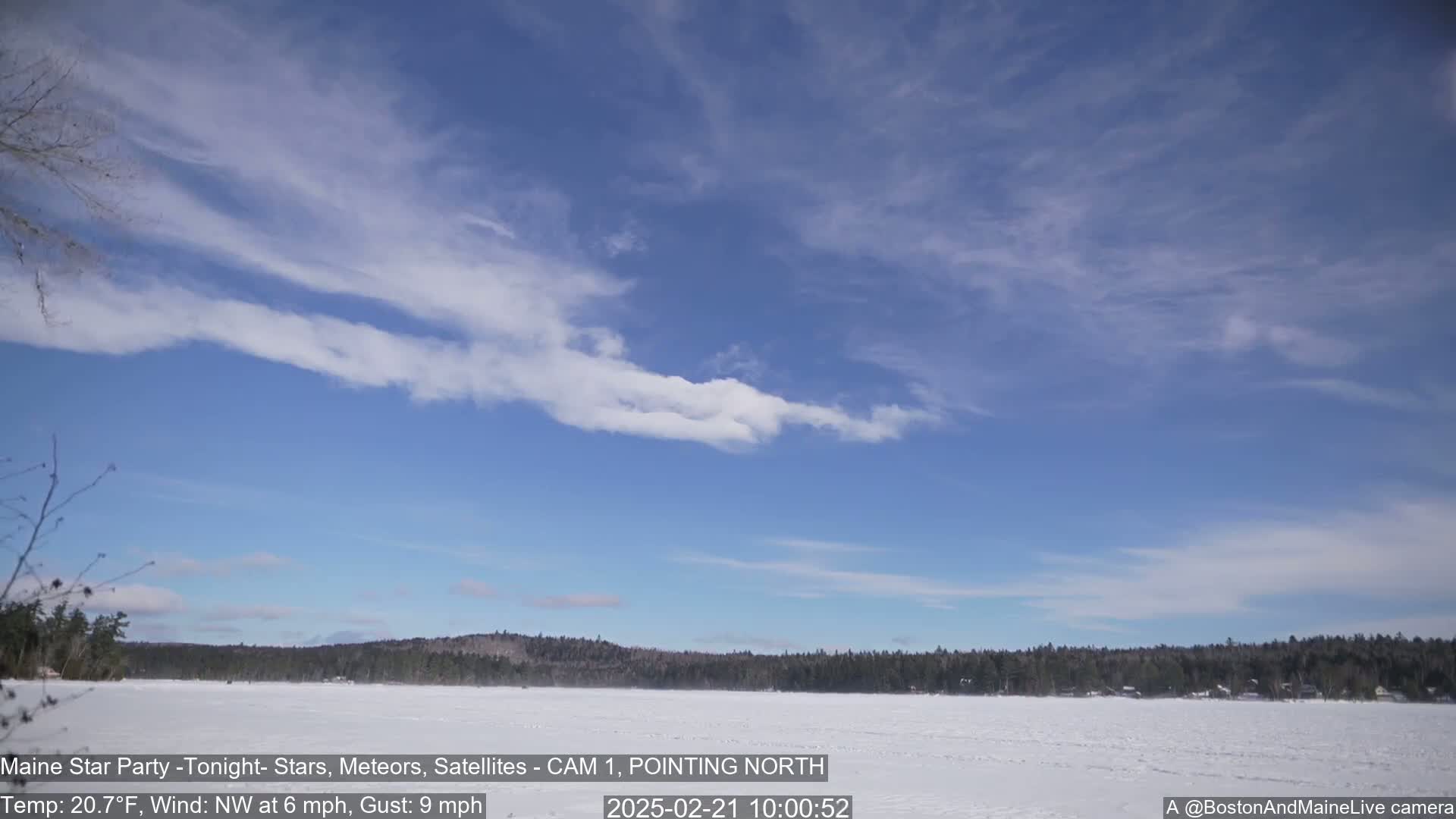 A mostly clear, blue sky with wispy clouds stretches over a frozen, snow-covered lake and a dark, forested shoreline.