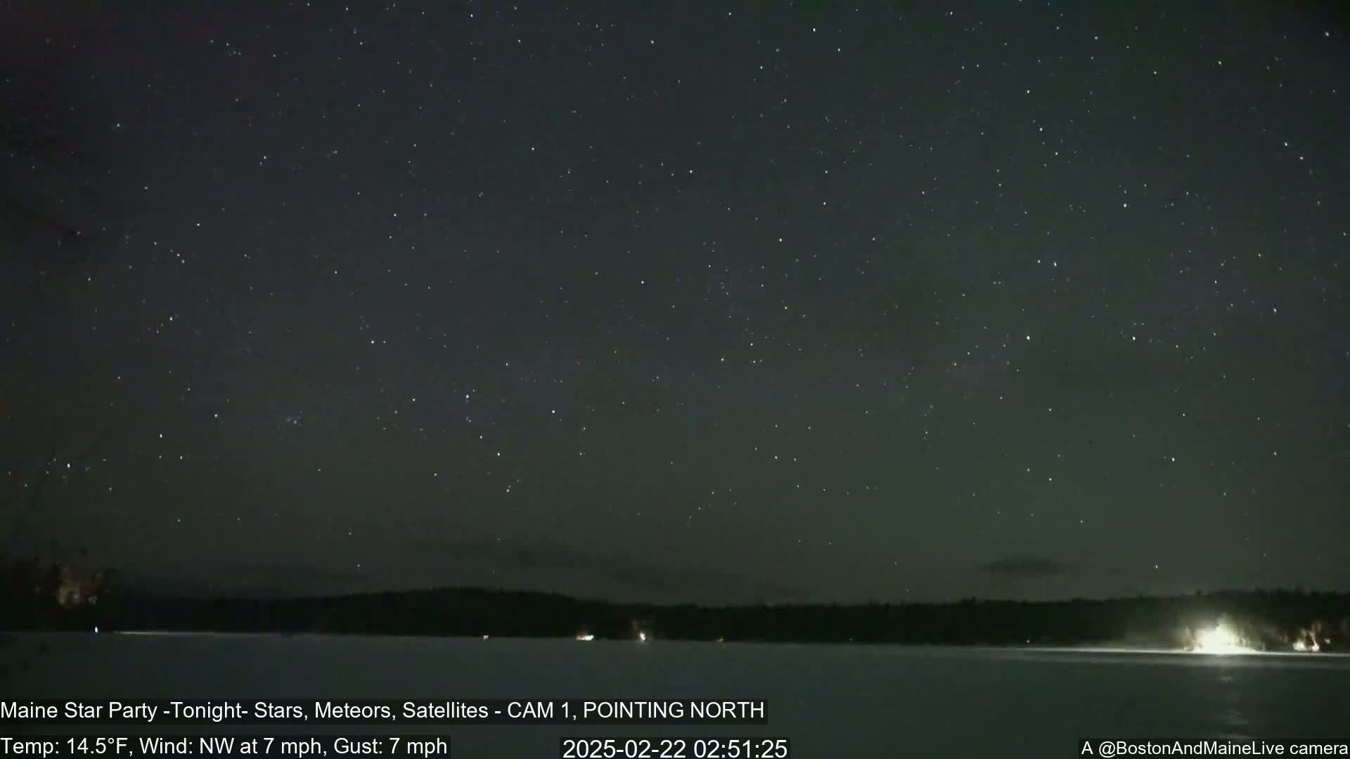 A nighttime long exposure shot shows a dark, clear sky full of stars over a snow-covered lake and a dark treeline, with a 7 mph northwest wind and 14.5°F temperature.