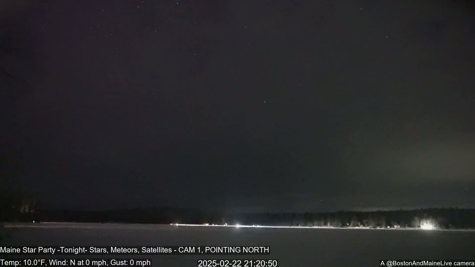 A nighttime long shot of a snow-covered landscape with a dark sky speckled with stars, and a faint line of lights visible on the horizon.