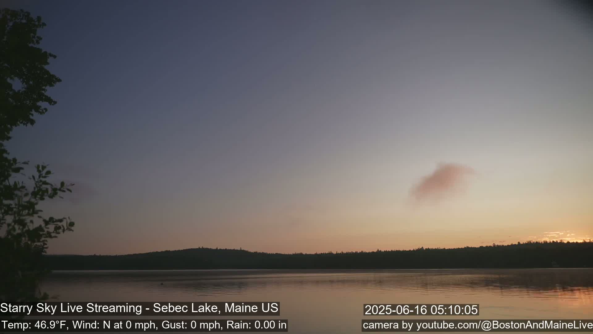 A calm lake at sunrise with a mostly clear sky and a single, light pink cloud is reflected on the still water.