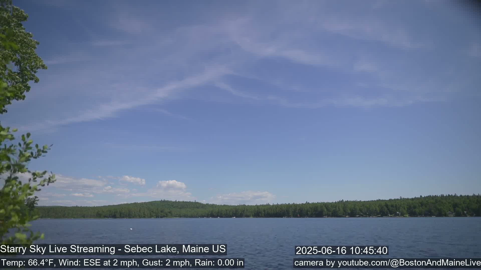 A calm lake with a tree-lined shore is visible under a mostly clear, light blue sky with a few wispy clouds.