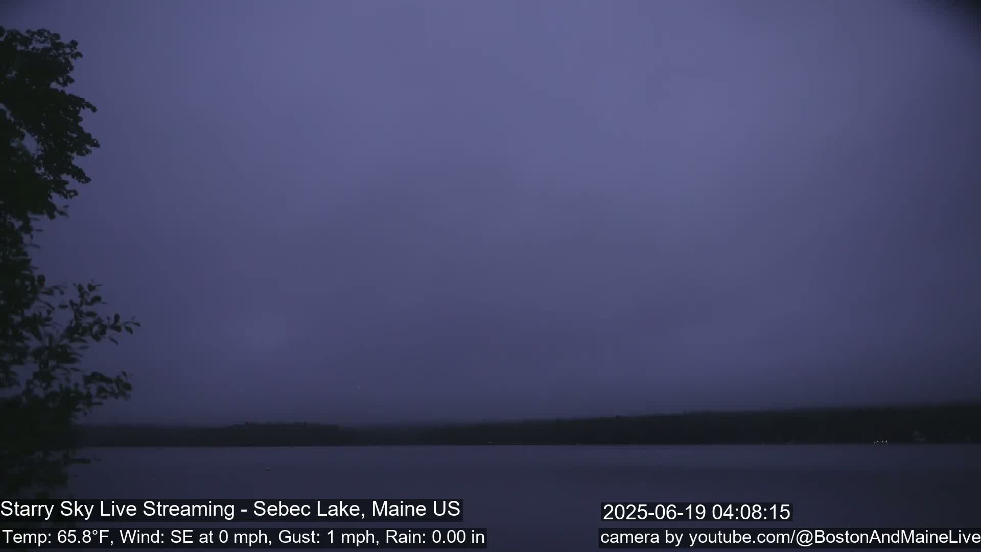 A dark, hazy, and still lake is seen under a dusky purple sky with a few dark trees to the left.
