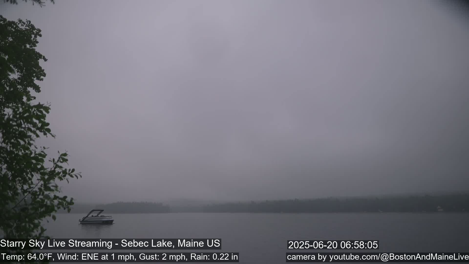 A boat sits on a calm lake shrouded in a thick fog under a gray sky, with light rain falling.