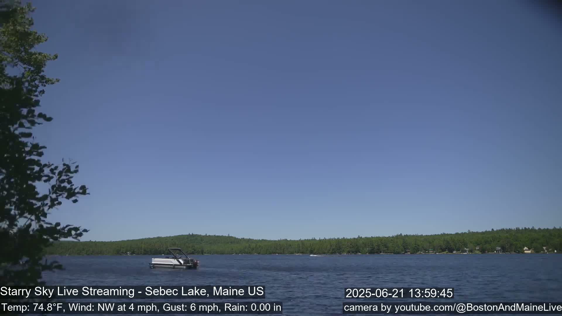 A pontoon boat sits on a calm lake under a clear blue sky, with a forested shoreline in the background.