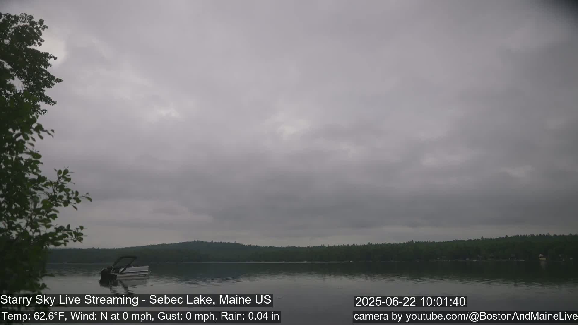 A pontoon boat sits on a calm lake under an overcast sky, with light rain.
