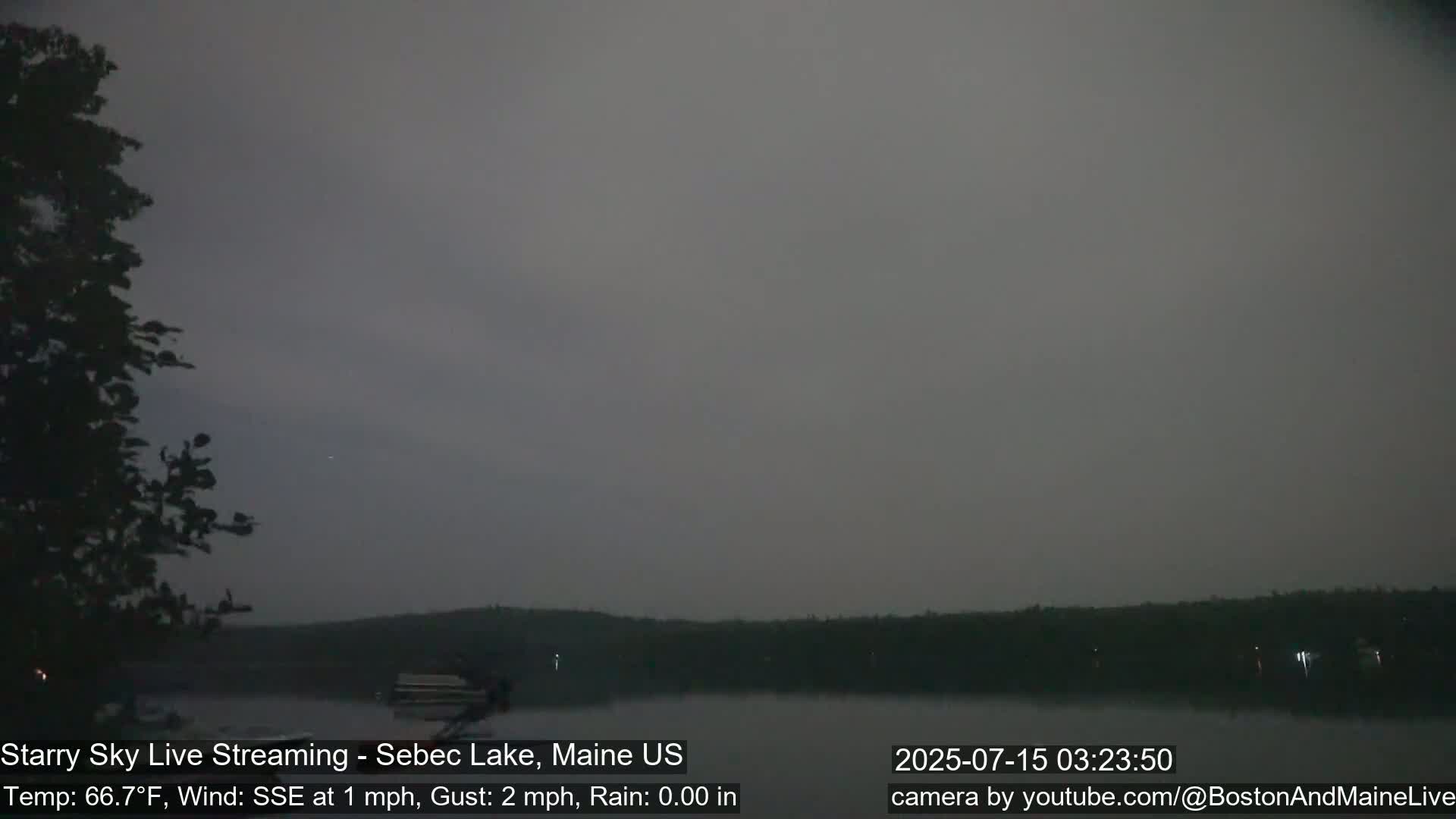 A dark, overcast night scene shows a calm lake with a treeline in the background, under a sky with a hint of light.